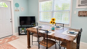 A dining area with a wooden table, chairs, a vase with sunflowers, and a television in the background. - 5 O'Clock Somewhere getaway-Walk/Bike to Beach,Shops ,Fenced private doggie area (Naples)