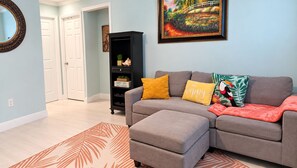 A living room with a grey sectional sofa, a black entertainment unit, and a colorful painting on the wall. - 5 O'Clock Somewhere getaway-Walk/Bike to Beach,Shops ,Fenced private doggie area (Naples)