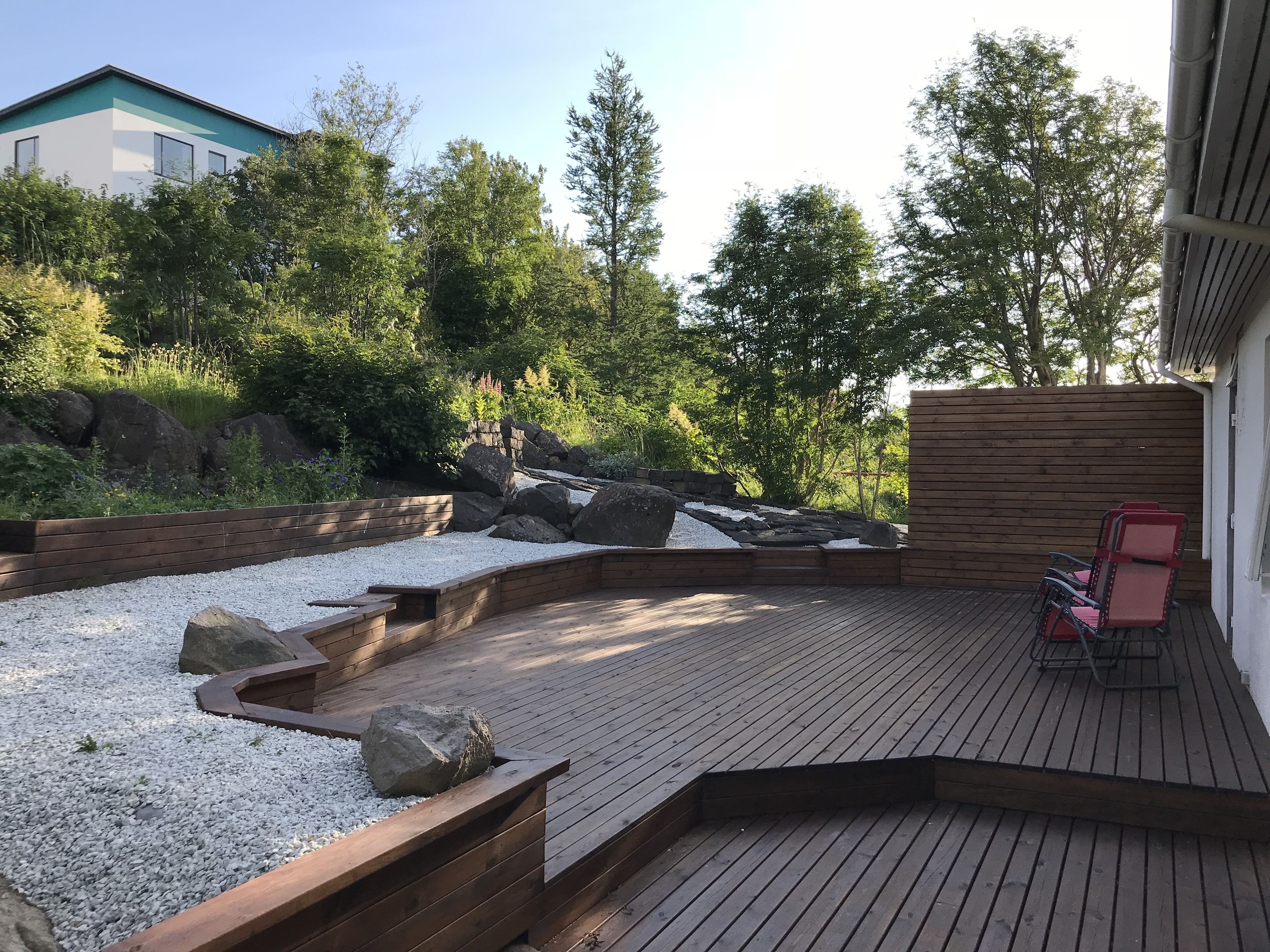 Eine Holzterrasse mit Sitzgelegenheit, umgeben von einem angelegten Garten mit Steinen und Kies.