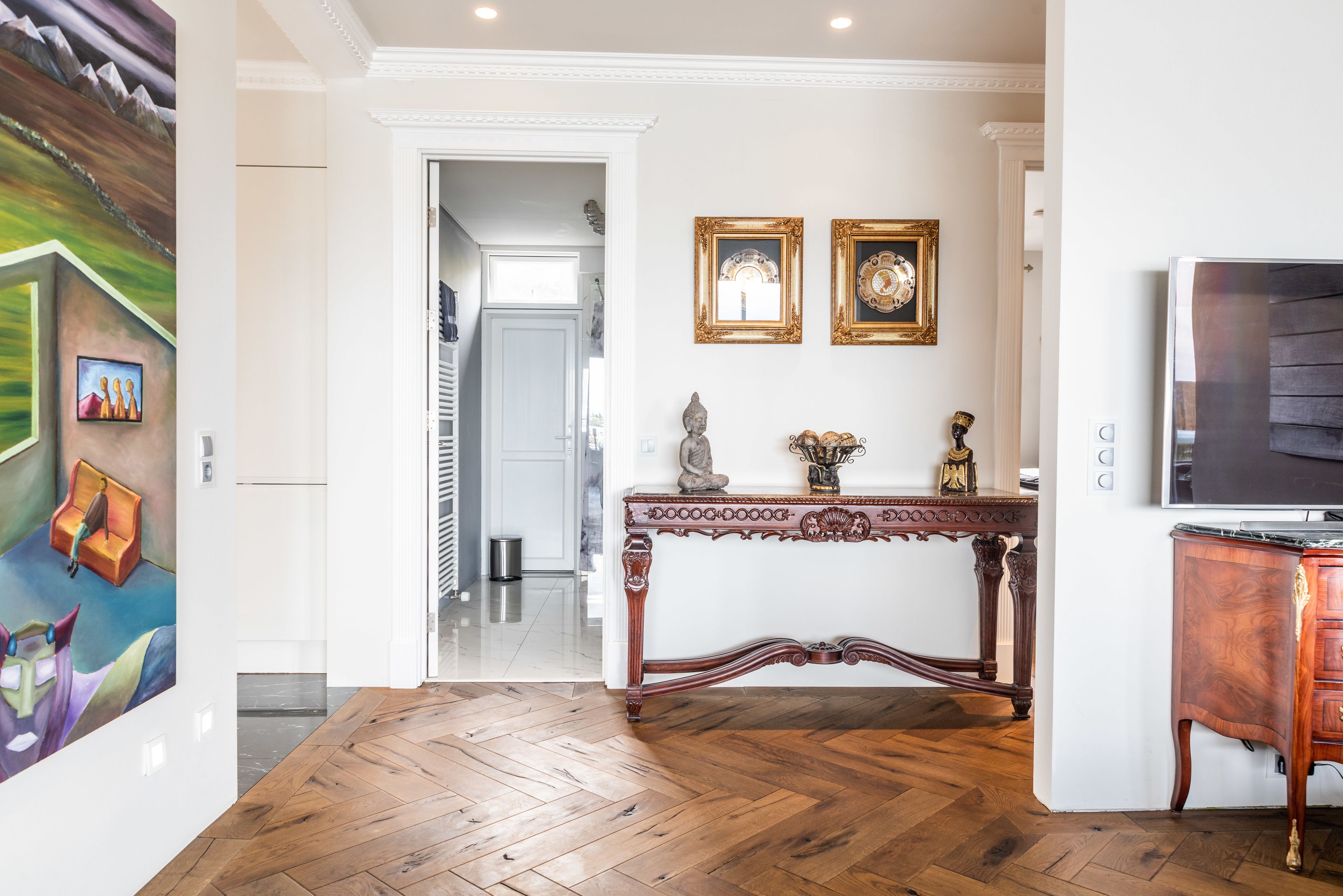 Un'ampia zona giorno con pavimento in parquet a spina di pesce, un grande televisore a schermo piatto, un tavolo consolle in legno con oggetti decorativi e un quadro appeso al muro.