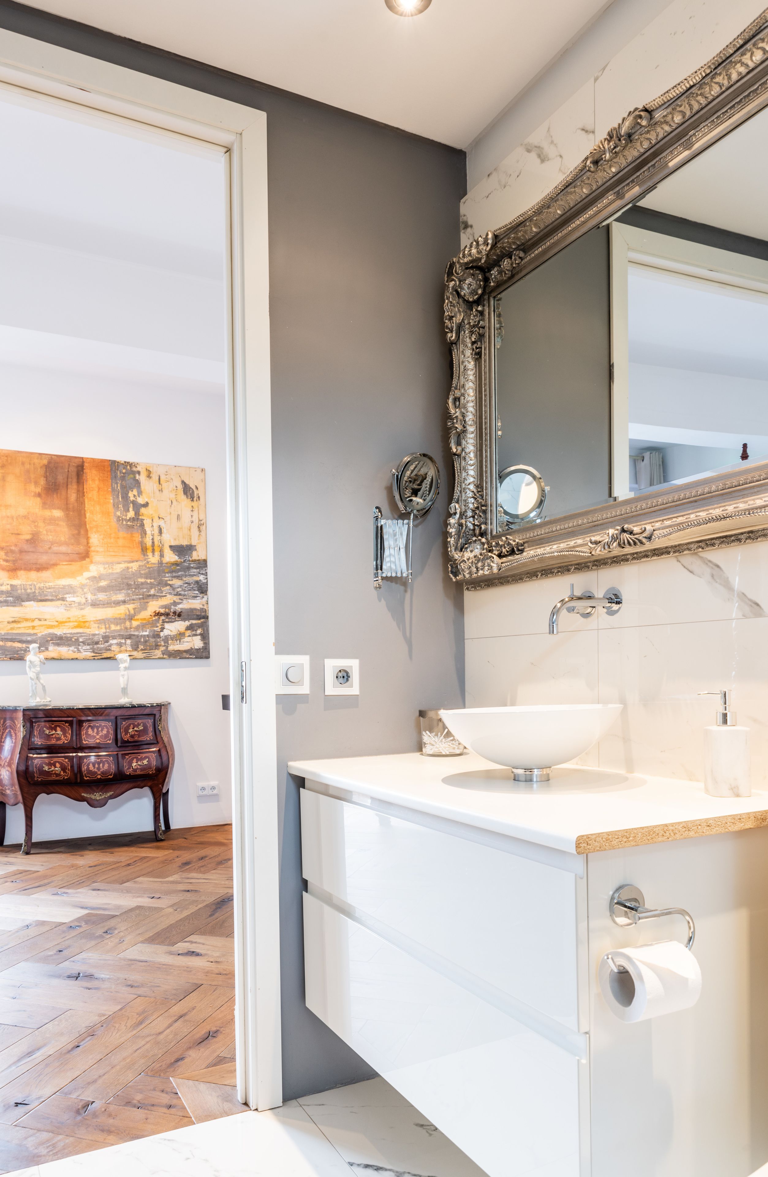 Un baño con un lavabo blanco, un espejo grande y decorativo, y suelo de madera.