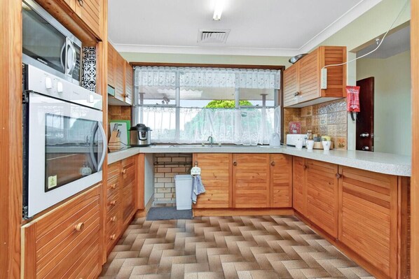 Una cocina compacta con armarios de madera, horno empotrado, microondas y una ventana con cortinas de encaje.
