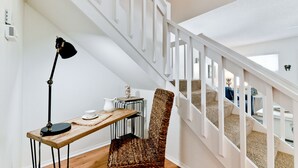 Een trap met witte leuningen, een houten tafel met een lamp, een geweven stoel, en een klein tafeltje met een kruik en beker.