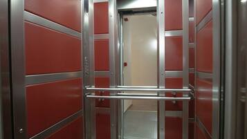An elevator with metallic doors and a handrail, featuring a red wall and a speckled floor.