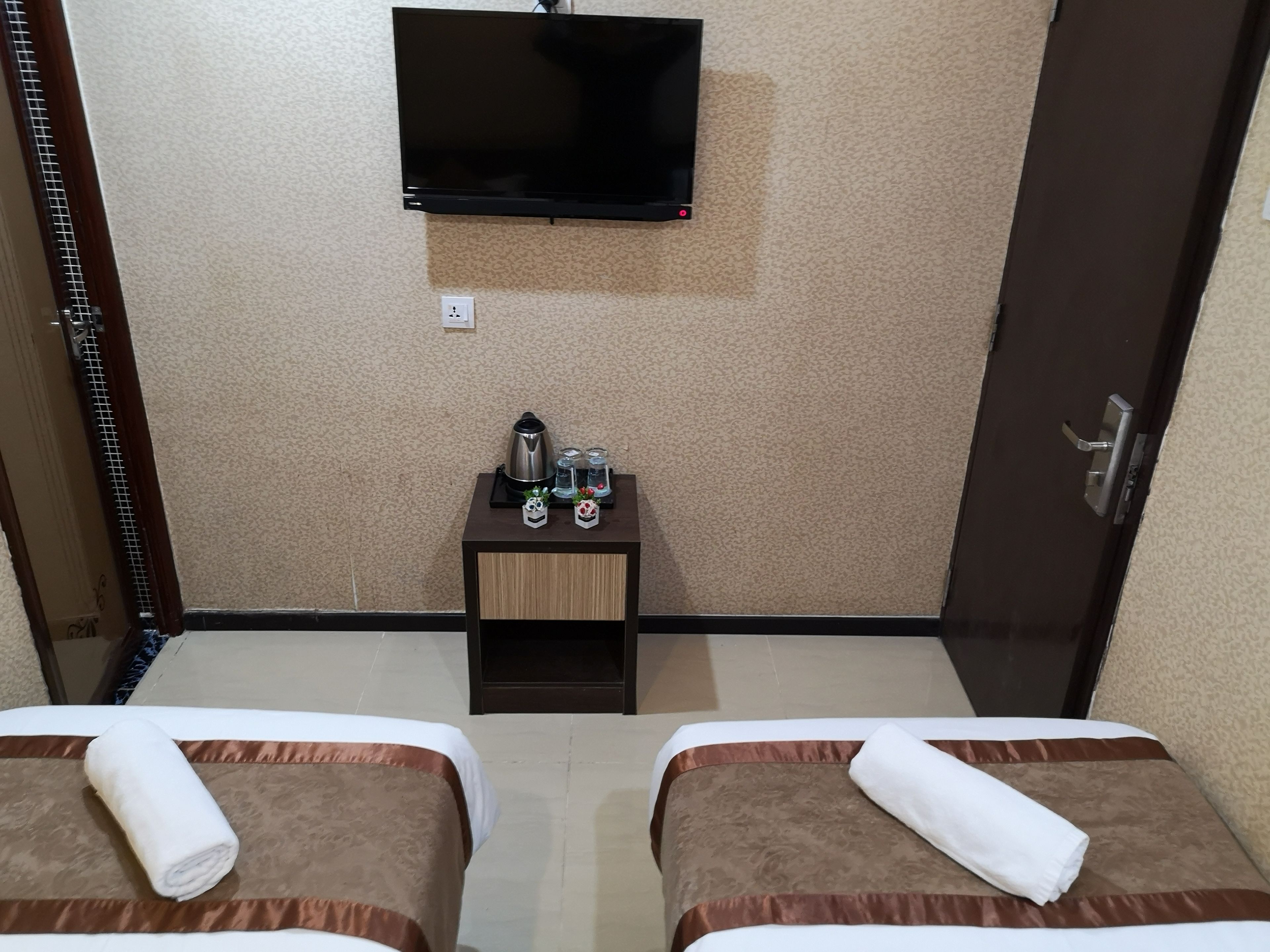 A hotel room with two beds, a TV mounted on the wall, a small table with a kettle and glasses, and a door with a handle.
