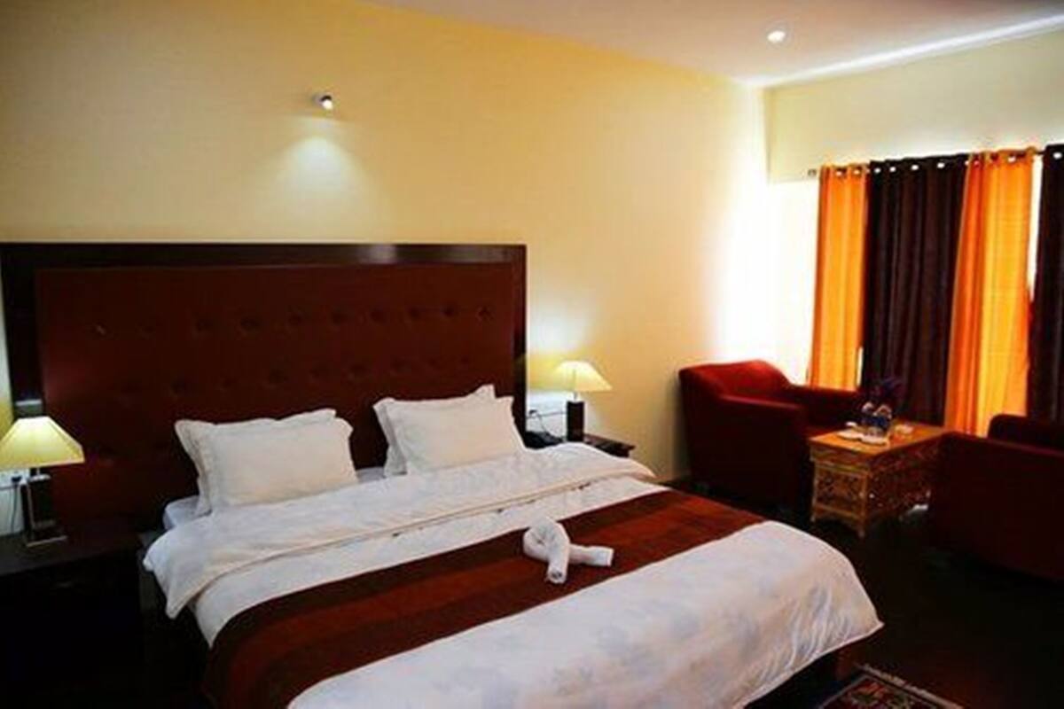 Habitación de hotel con una cama grande, dos mesitas de noche con lámparas, un sillón rojo y una ventana con cortinas naranjas.