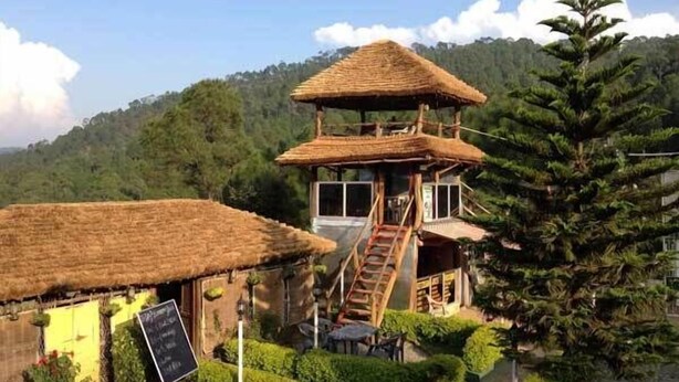 A rustic wooden structure with a thatched roof, a staircase leading to a balcony, and a small garden area with neatly trimmed bushes. - Uttarakhand Resort (Somashwar)