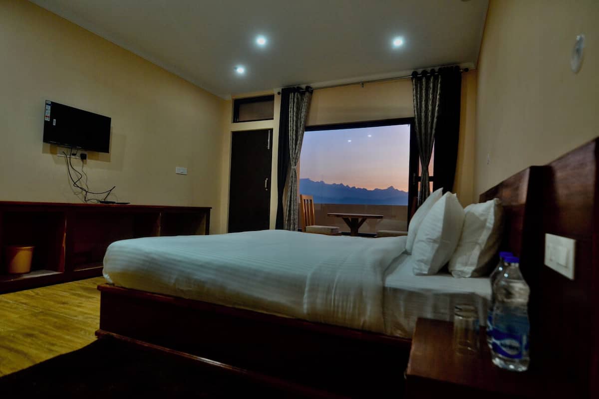 Habitación de hotel con cama, televisor montado en la pared, ventana con cortinas y vistas a montañas.