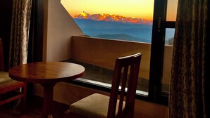 A room with a wooden table and a cushioned bench, a window with a view of a sunset over mountains, and curtains on either side. - Uttarakhand Resort (Somashwar)