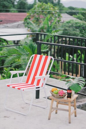 Ein rot-weiß gestreifter Liegestuhl auf einem Balkon, mit einem kleinen Holzschemel, der einen Obstkorb und eine Flasche trägt.