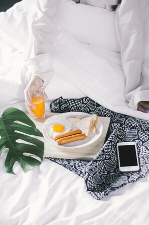 Ein ordentlich gemachtes Bett mit weißer Decke, ein Teller mit Eiern, Toast und Würstchen, ein Glas Orangensaft und ein Smartphone auf einem gemusterten Tuch.