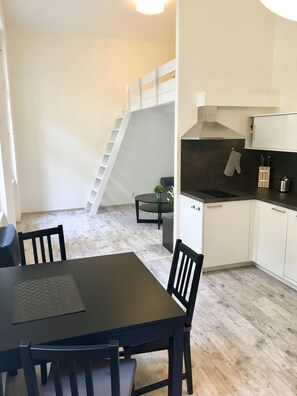 Moderni keittiö, jossa on valkoiset kaapit, musta työtaso ja sisäänrakennettu liesi. Keittiössä on myös ruokailutila, jossa on pöytä ja tuolit, sekä portaat, jotka johtavat ylemmälle tasolle.