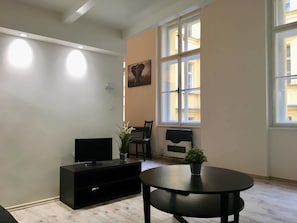 A room with a black entertainment unit, a round table with a potted plant, a chair, and a window with a view of buildings. - Hybernska (Prague)