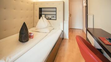 A hotel room with a bed, a bedside table, a red bench, a wall-mounted shelf with a telephone, and a door leading to another room.