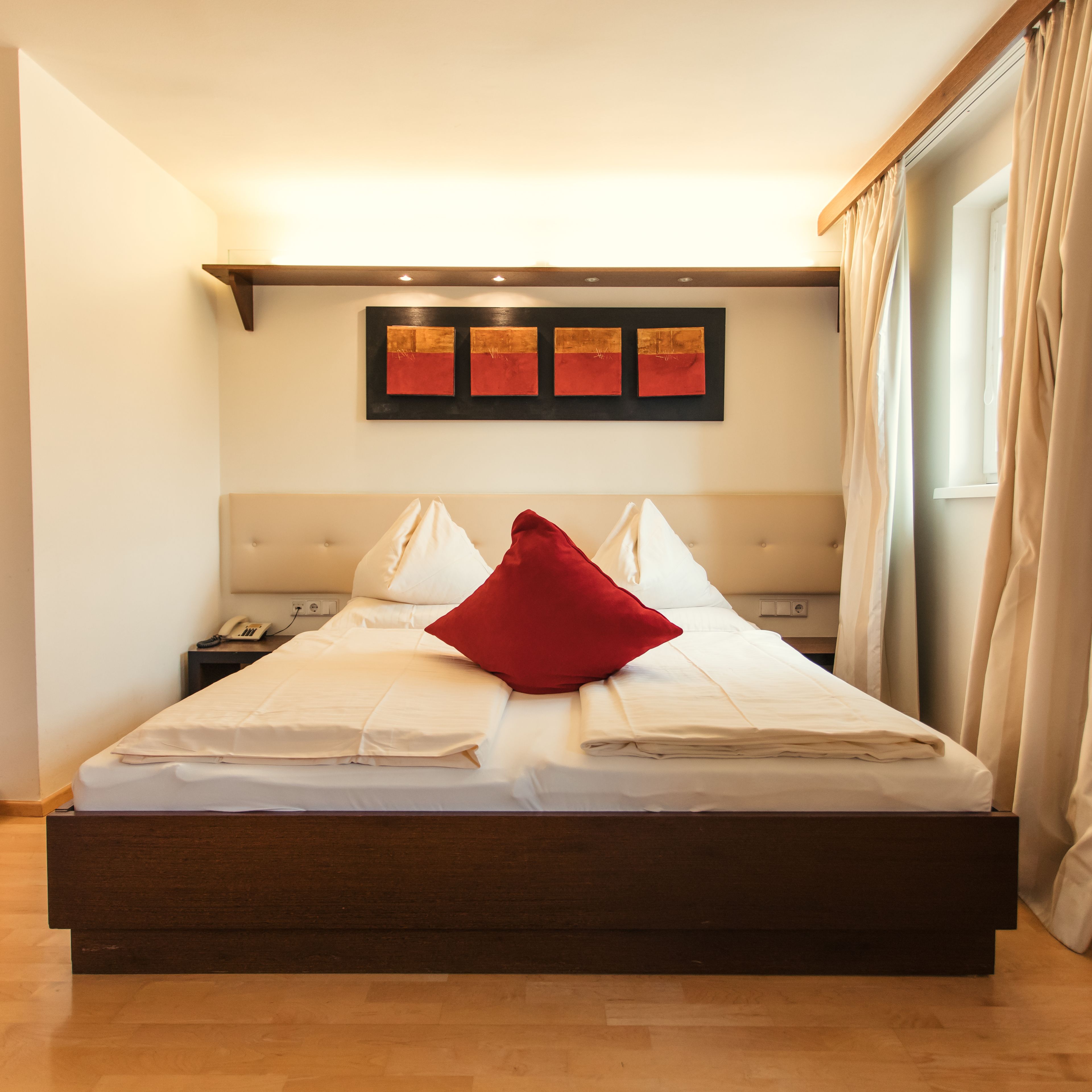 A neatly made bed with white linens and a red pillow, a bedside table with a phone, and a wall with three abstract paintings.