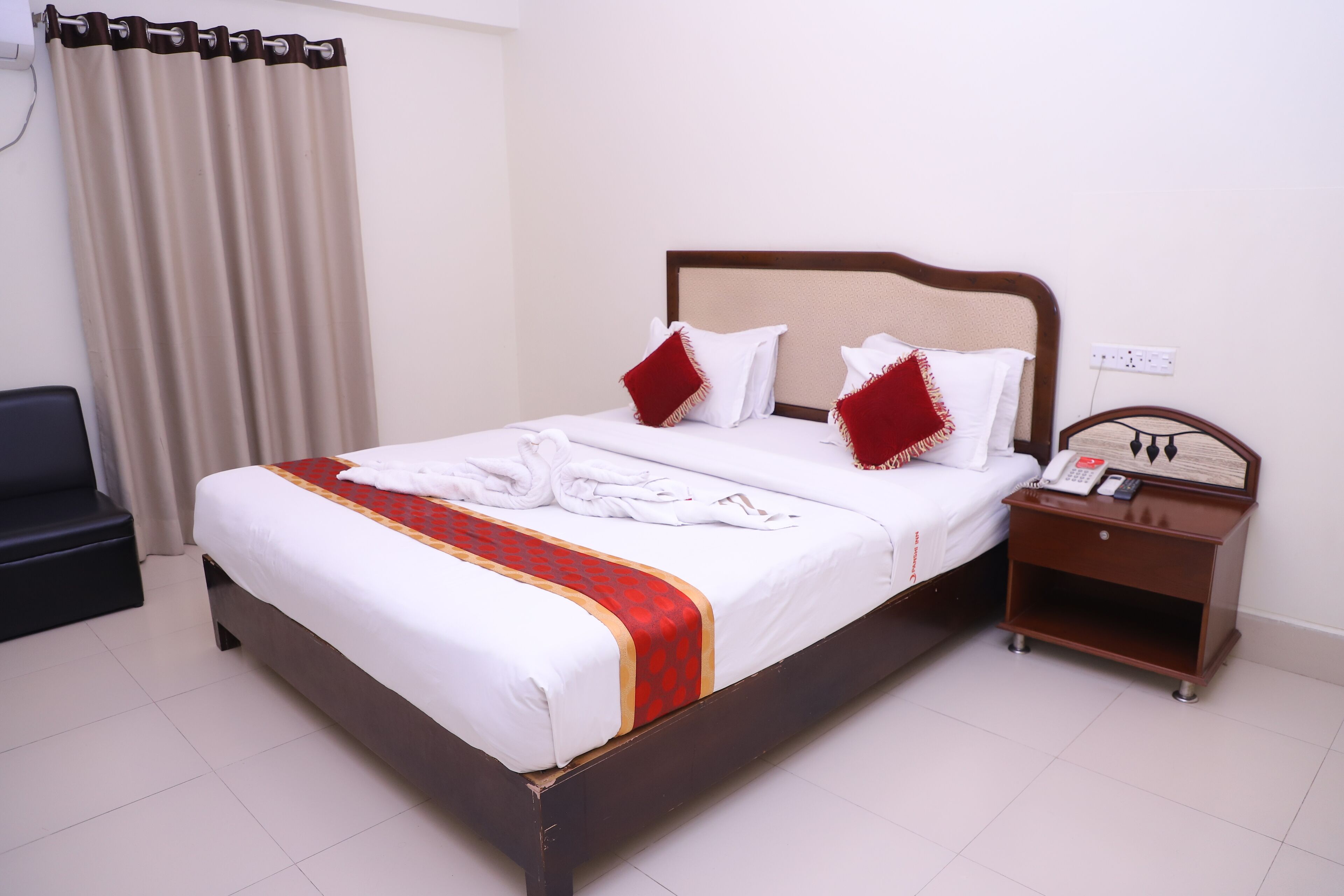 Een netjes opgemaakt bed met een rood-witte spreideken, twee kussens en een houten nachtkastje met een telefoon en afstandsbediening.