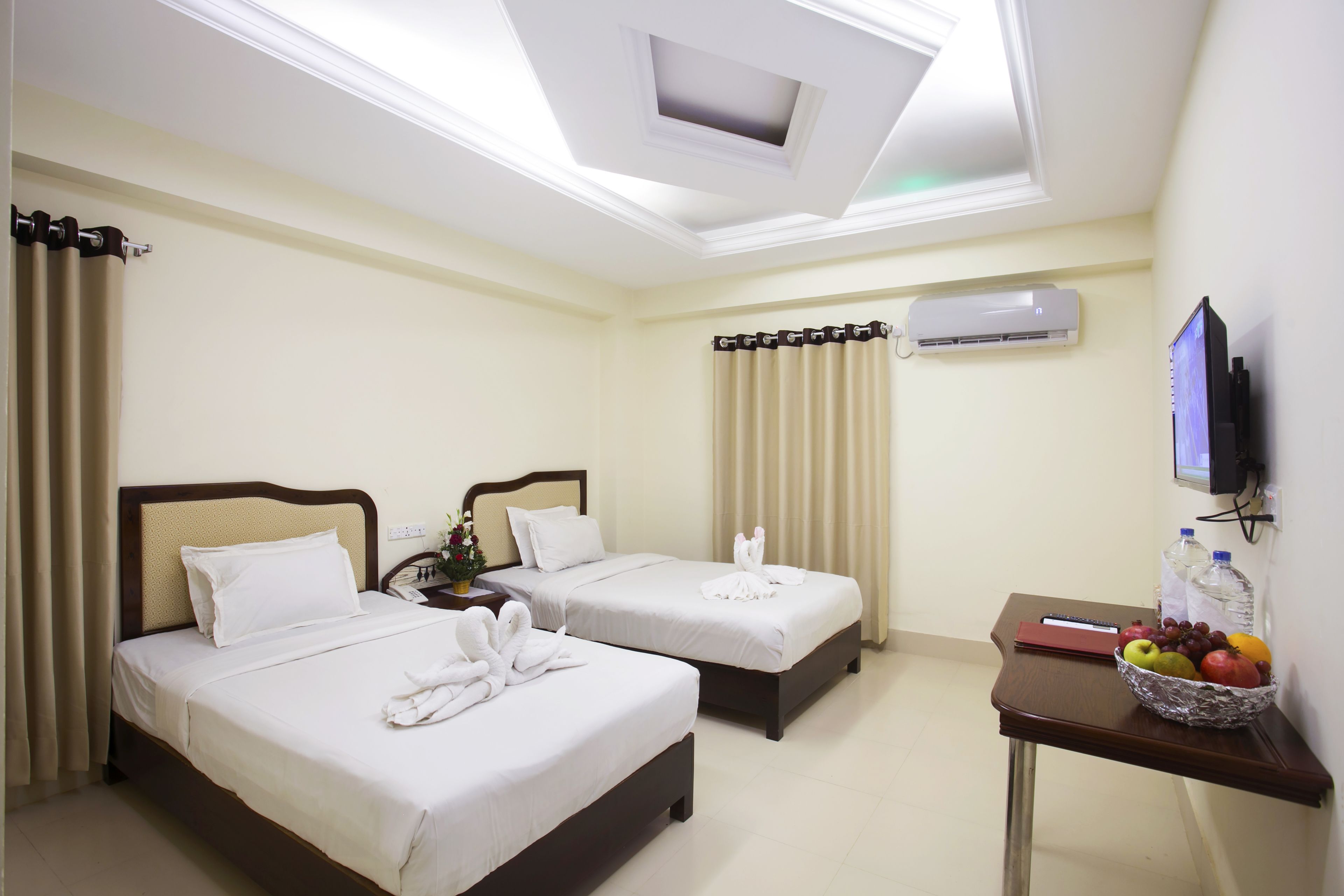 A hotel room with two beds, a desk with a fruit bowl, a TV, and an air conditioning unit.