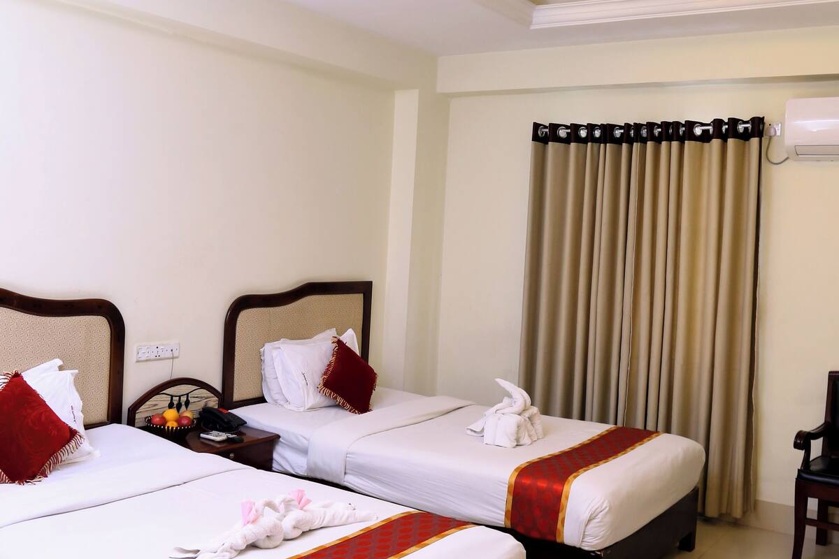 Quarto de hotel com duas camas, uma mesa de cabeceira, escrivaninha e janela com cortinas.