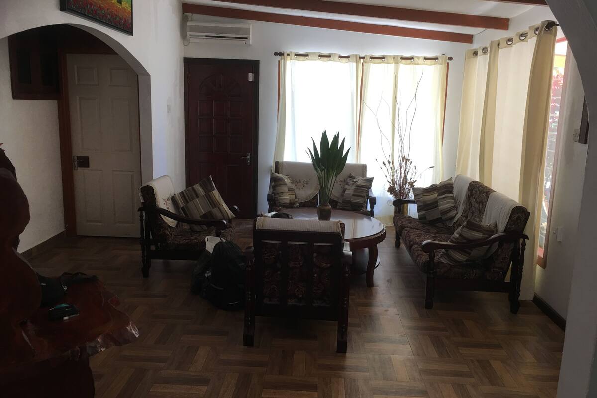 A living room with a wooden herringbone floor, a round coffee table, two sofas, and a dining table. There is a painting on the wall, a door, and a window with curtains.