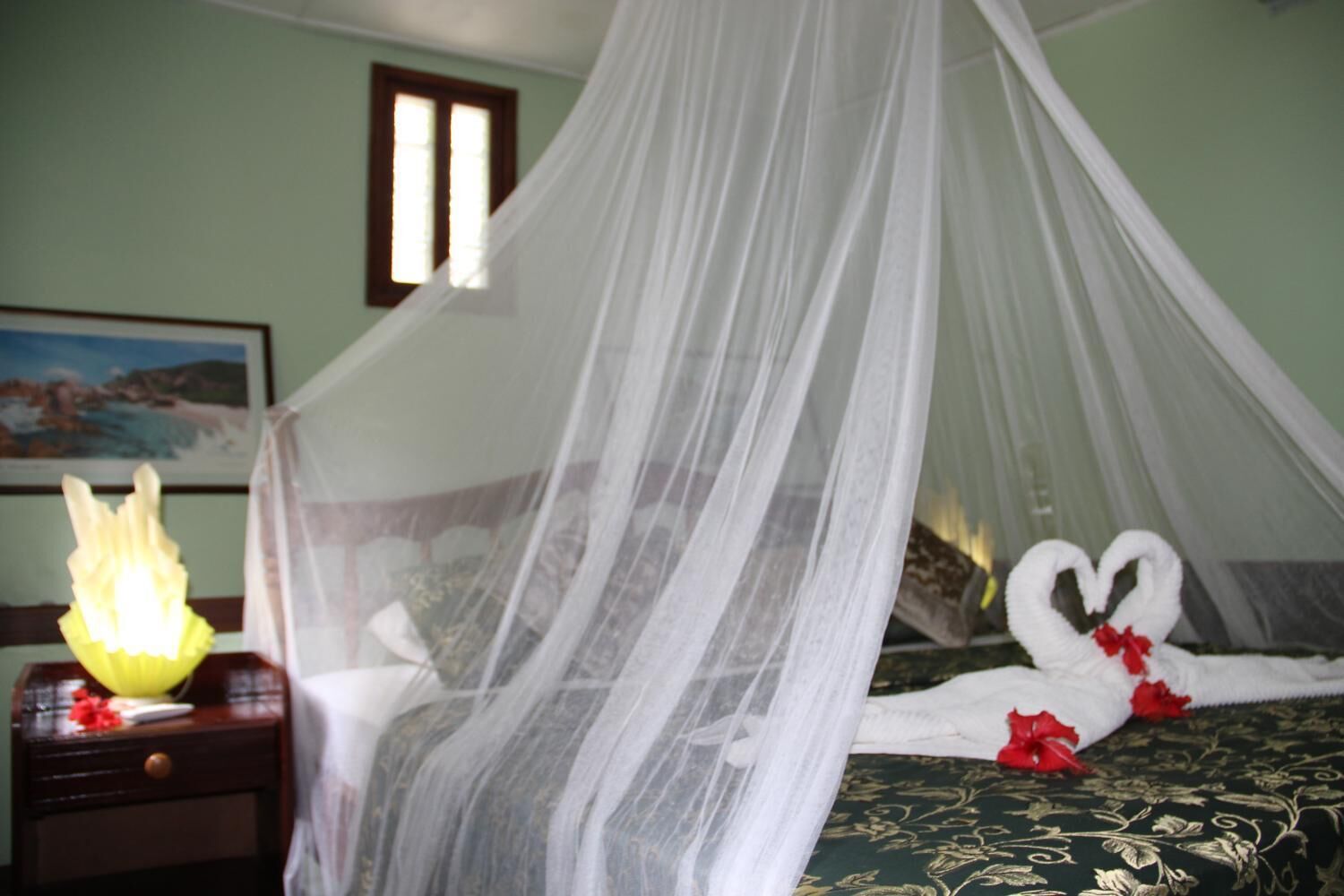 Una habitación con una cama bajo un dosel blanco, una mesita de noche con una lámpara y una colcha con una disposición de toallas en forma de corazón.