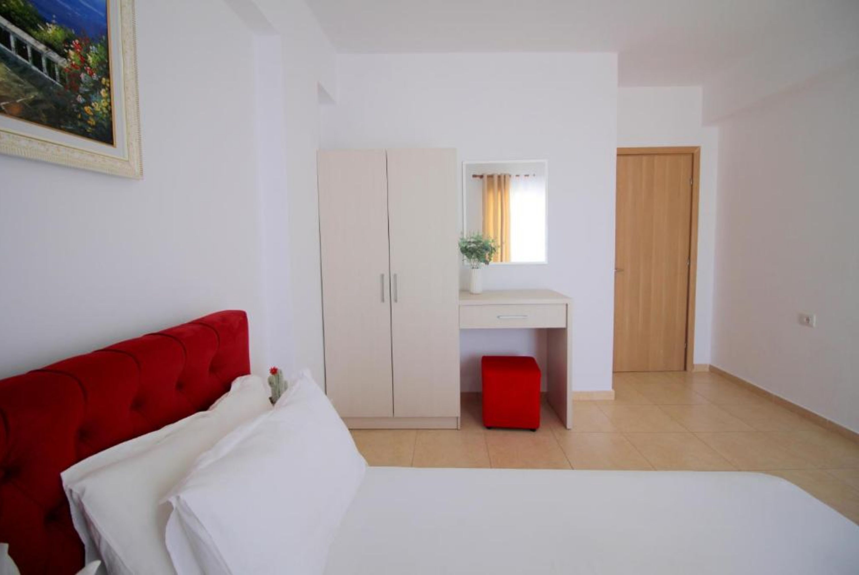 A bedroom with a bed, a red headboard, a white wardrobe, a desk with a mirror, and a window with curtains.