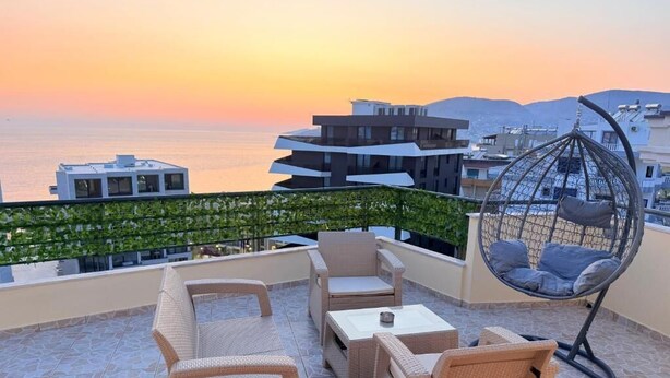 A rooftop terrace with wicker furniture, a hanging chair, and a view of the sea and buildings at sunset. - AR Apartments by Saranda Hotel (Sarandë)