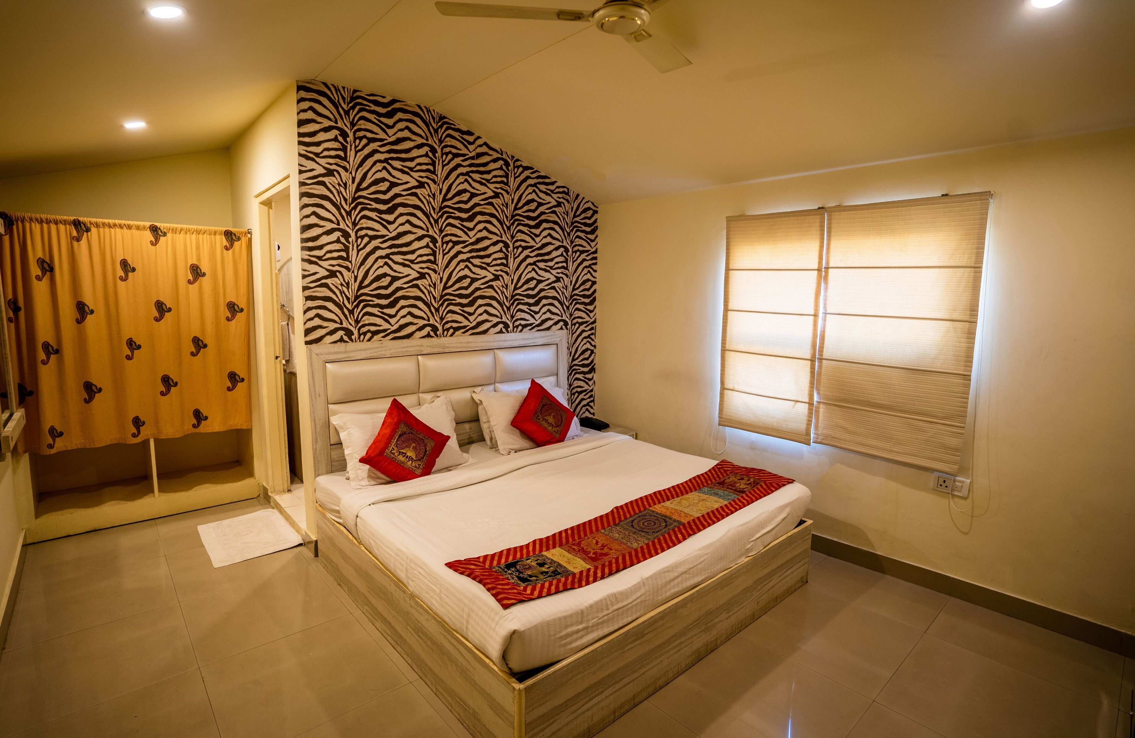A bedroom with a zebra-patterned headboard, a bed with white linens and red pillows, a window with blinds, and a ceiling fan.