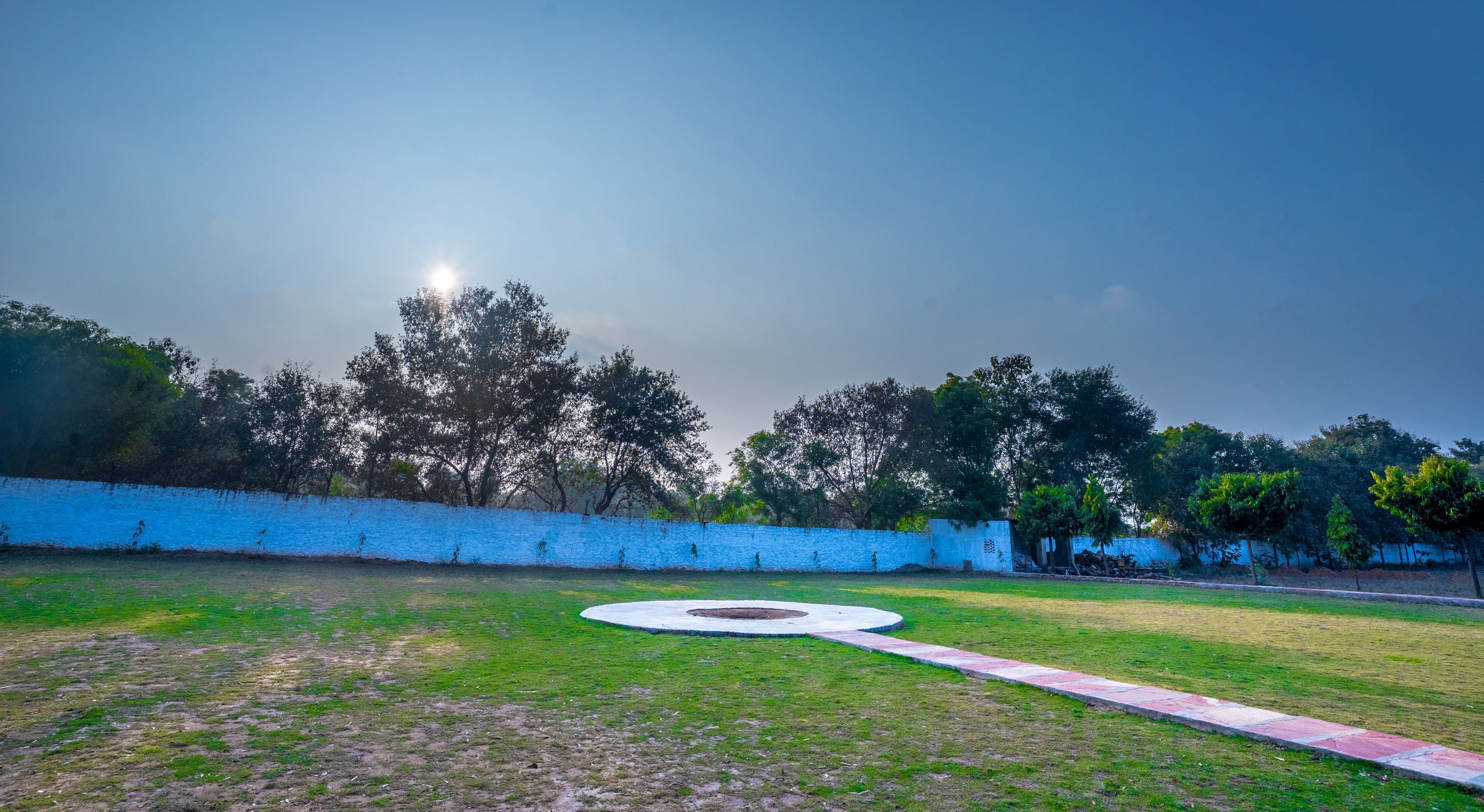 A grassy park area with a circular concrete feature, a blue wall, and trees in the background.