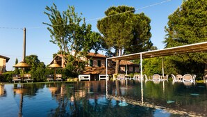 An infinity pool with lounge chairs and umbrellas, surrounded by trees and a building with a tiled roof. - Trere Agriturismo (Faenza)