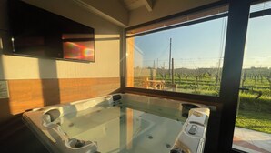 A spa room with a large hot tub, wooden paneling, a television, and a view of a vineyard through a large window. - Trere Agriturismo (Faenza)
