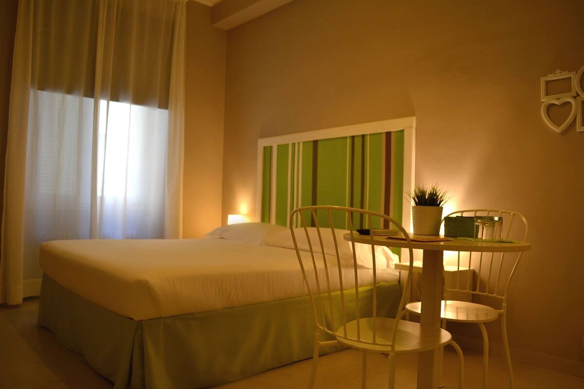 A hotel room with a bed, a small round table with chairs, a potted plant, and a wall-mounted decoration.
