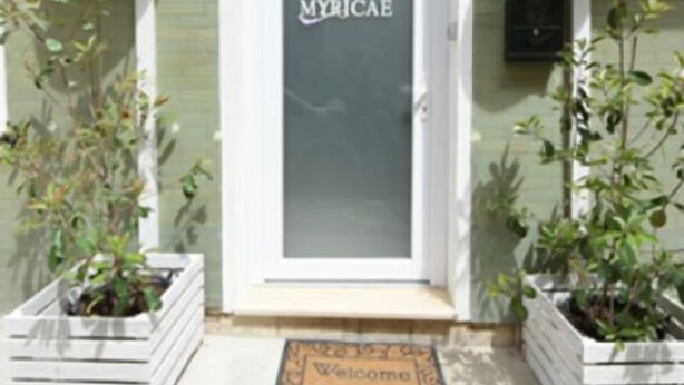 Une entrée d'immeuble avec une porte blanche, une enseigne indiquant « MYRICAe » et deux plantes en pot de chaque côté.
