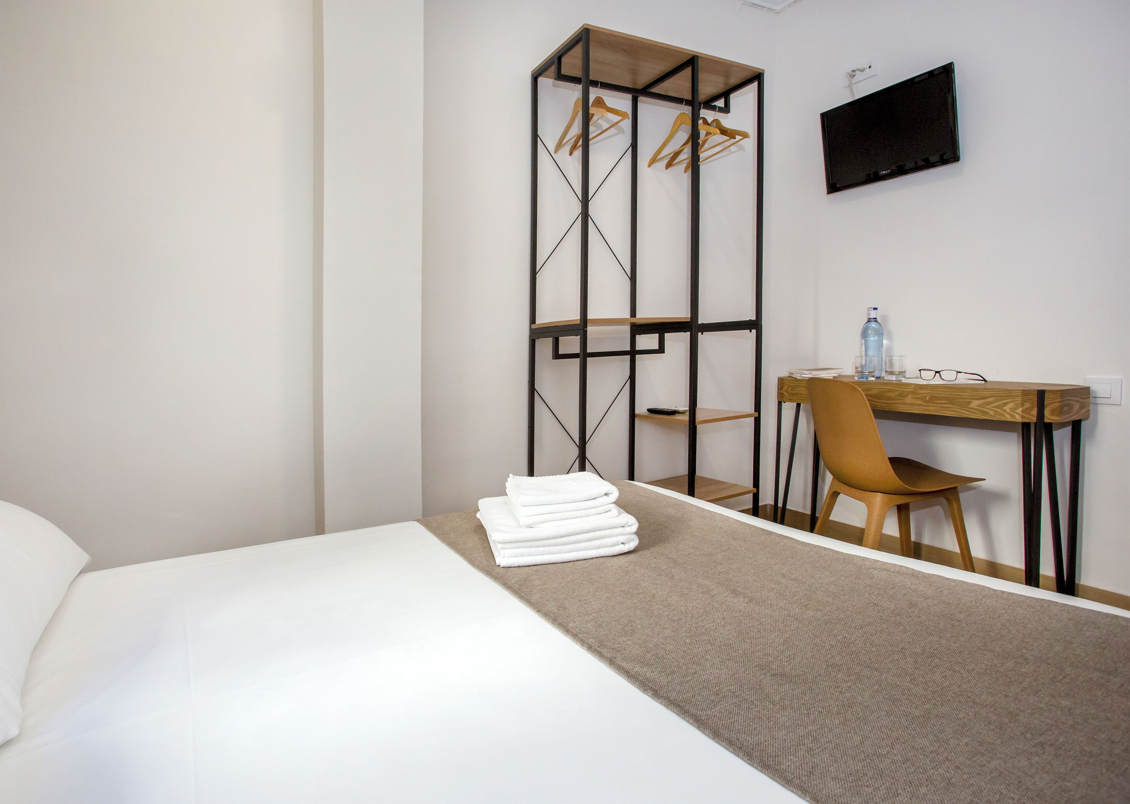 A hotel room with a bed, a desk with a chair, a TV, and a wardrobe with hanging clothes.