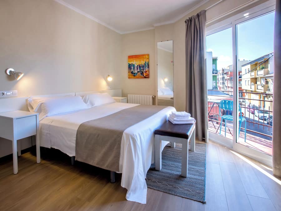 Una habitación de hotel con una cama, mesitas de noche, un banco, un ventanal con vistas a edificios y un cuadro en la pared.