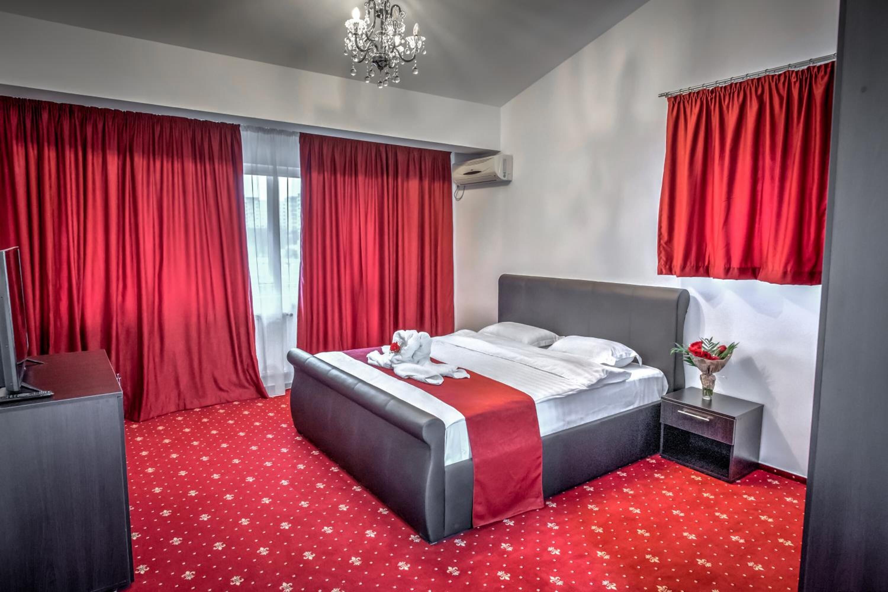 A hotel room with a large bed, red curtains, a chandelier, a small bedside table with a vase of flowers, and a red carpet.