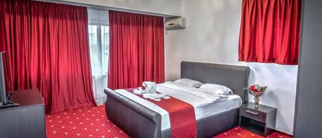 A hotel room with a large bed, red curtains, a chandelier, a small bedside table with a vase of flowers, and a red carpet.