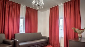 Una habitación con un sofá gris, un sillón pequeño y un escritorio de madera con un jarrón de flores. Se ven cortinas rojas y un candelabro.
