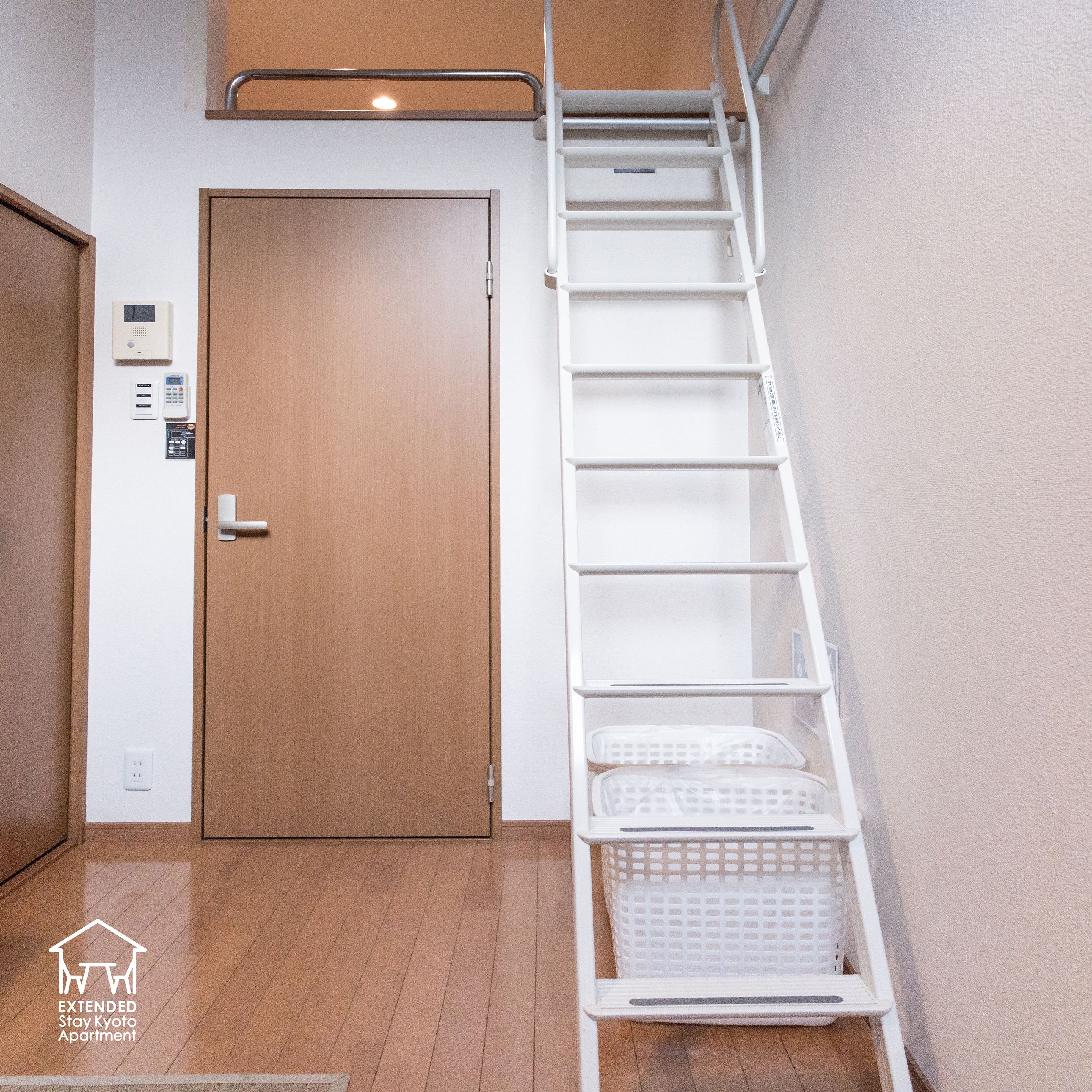 A modern apartment with a wooden door, a white wall-mounted security system, a white metal ladder, and a small white basket.