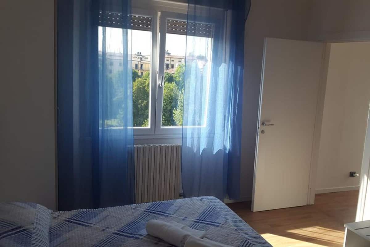 Ein Schlafzimmer mit einem Bett, einem Fenster mit blauen Vorhängen, einem Heizkörper und einer Tür zu einem anderen Raum.
