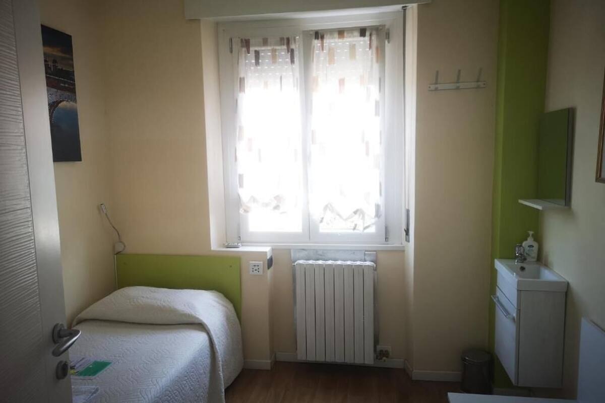 Ein kleines, helles Schlafzimmer mit einem Einzelbett, einem Heizkörper, einem Fenster mit Vorhängen, einem kleinen Schreibtisch und einem Spiegel.