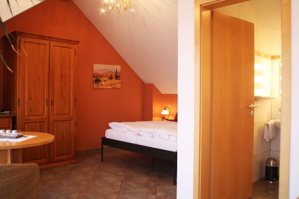 A bedroom with a bed, a wooden wardrobe, a painting, and a small table with a lamp.