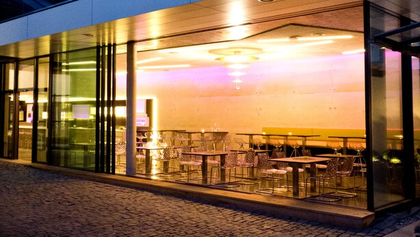 A modern building with a glass facade, outdoor seating area, and a visible interior space with tables and chairs. - Porta Vinea (Ruppertsberg)