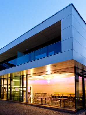 A modern building with a glass facade, outdoor seating area, and a visible interior space with tables and chairs. - Porta Vinea (Ruppertsberg)