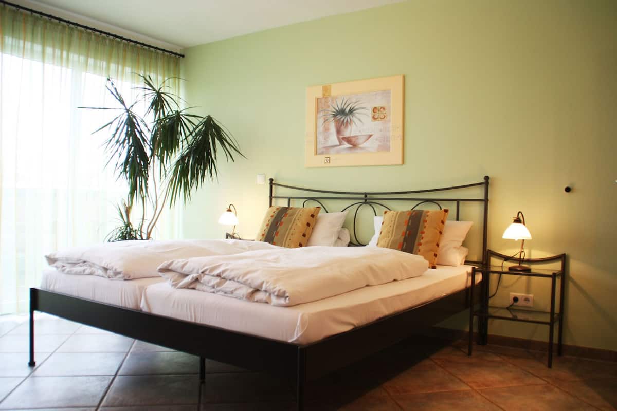 A neatly made bed with white linens, a large potted plant to the left, and a framed artwork on the wall.
