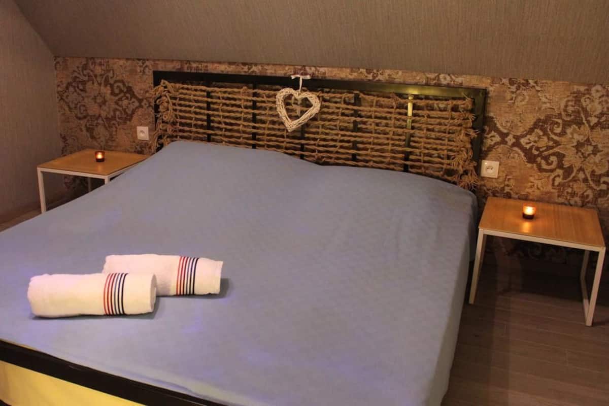 A neatly made bed with a blue bedspread, two bedside tables with candles, and a decorative heart hanging on the headboard.