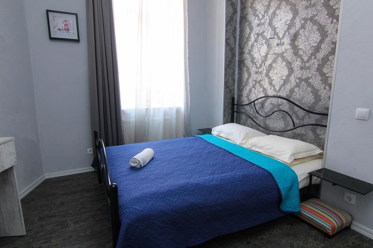 A neatly made bed with a blue bedspread and white pillows, a small bedside table, a striped cushion, and a patterned wallpaper on the wall.