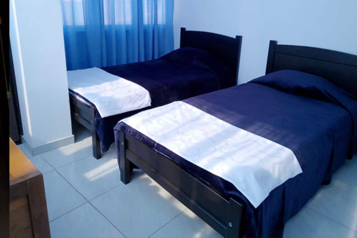 Una habitación con dos camas individuales, una mesita de noche de madera y una ventana con cortinas azules.