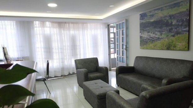 Una sala de estar moderna con un sofá gris, un reposapiés a juego y una mesa de centro. Hay un ventanal con cortinas translúcidas, un cuadro en la pared y una puerta que da al exterior.
