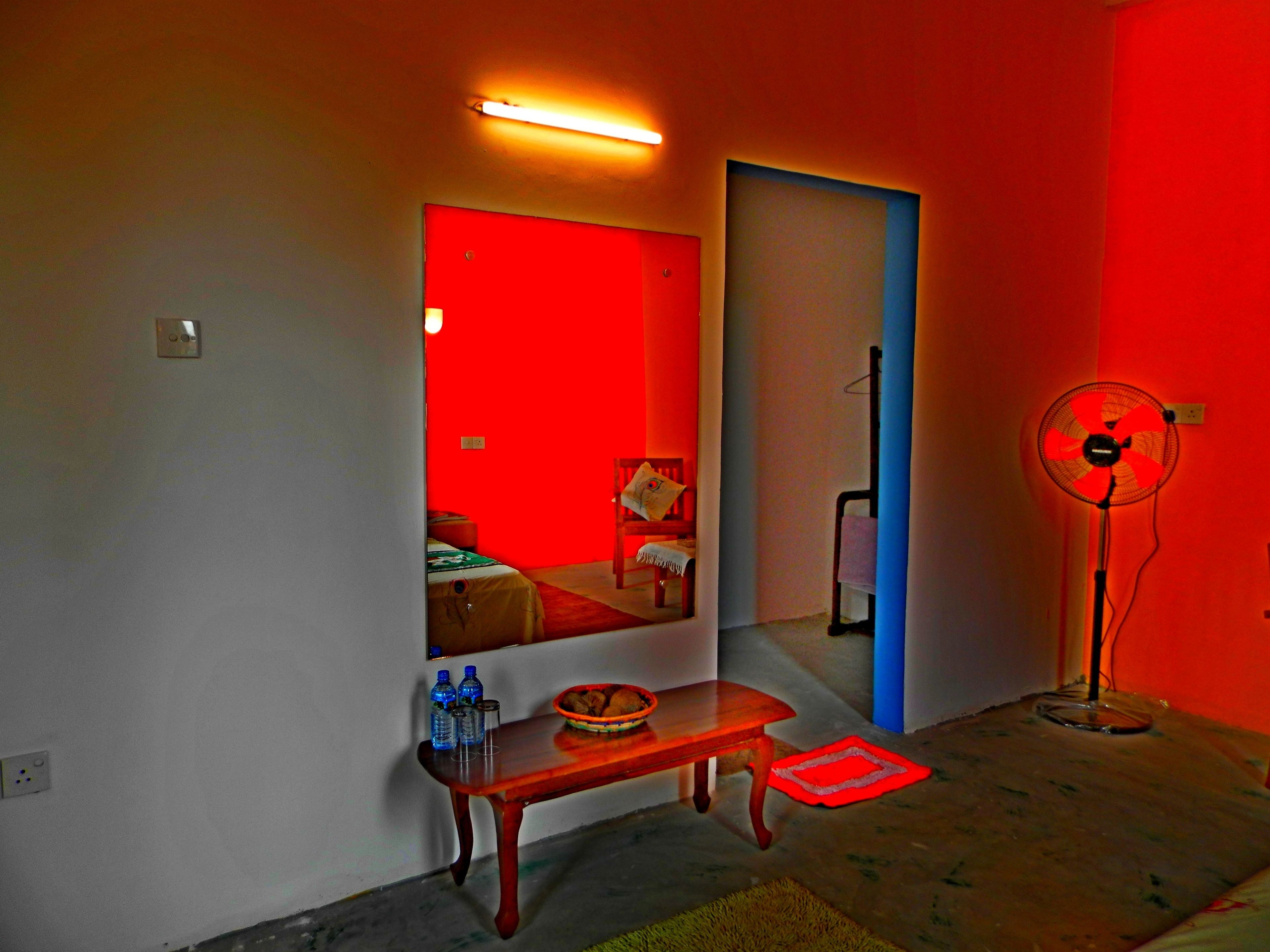 Una stanza con una parete rossa, uno specchio, una panca di legno, una ventola e un tavolino con bottiglie e un cesto.