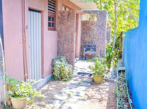 A narrow pathway leading to a building with a door and window, surrounded by potted plants and a blue wall.